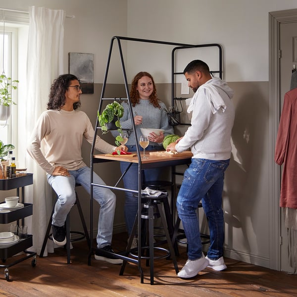 Three gather around modern black metal table, one stands, one holds food, casual setting.