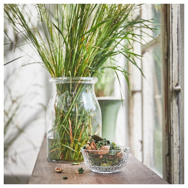 A clear glass vase with green plants sits on a wooden shelf next to a small bowl of green potpourri.