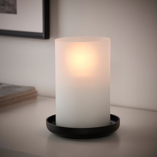 A frosted glass candle, lit and placed on a black round holder, emitting a warm glow.