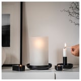 Black HEDERVÄRD candle holders with frosted glass, creating soft glow, arranged on a white surface with lit candles.