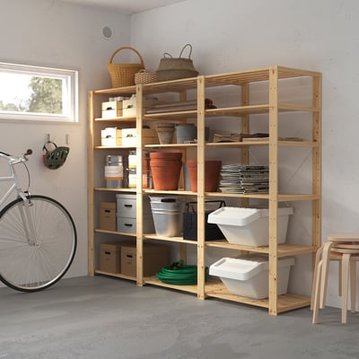 A HEJNE storage system made of light-colored wood with multiple shelves holding various items. Its larger than the bicycle and stool placed nearby.