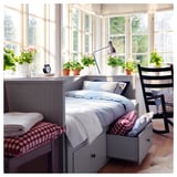 Bright bedroom with large windows, grey day-bed, red checkered pillows, black rocking chair, and potted plants.
