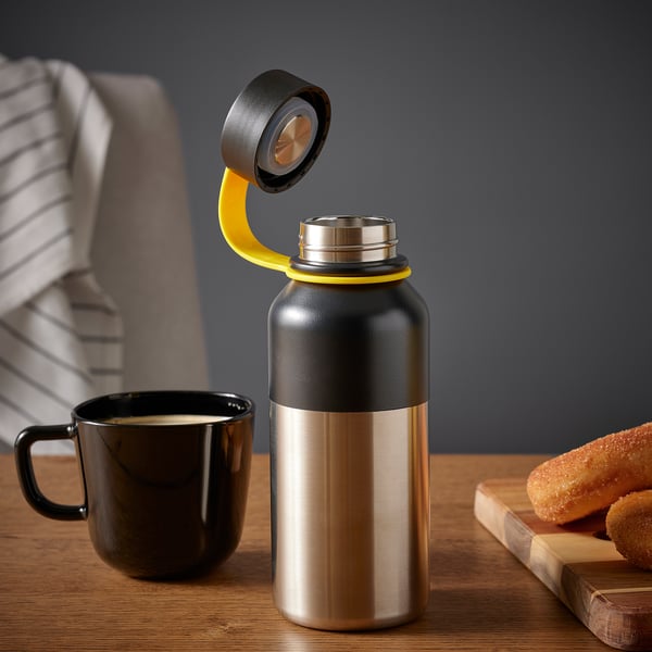 Thermal flask with yellow handle on wooden table, beside black coffee mug and pastry. Lid opens to reveal spout for pouring.