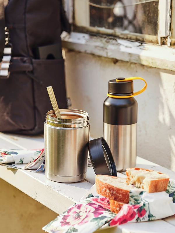 Silver insulated flask with black lid, yellow handle, on wood. Beside another flask and bread on cloth.