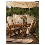 Outdoor setting with beige HÖGÖN parasol and wooden table; four chairs, cushions, flowers, and fruits on the table, surrounded by plants.