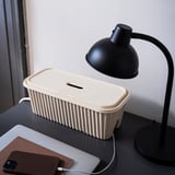 Desk with black lamp, honsnät beige cable box, brown notebook, and charging phone. Box hides cables neatly.