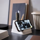 Small beige HÖNSNÄT mobile phone & pen stand, angled phone display, near books & bin.