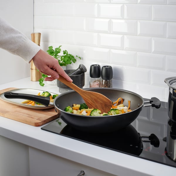 A person cooks colorful veggies in a black pan on a sleek stove. A wooden spatula stirs them, while a cutting board with fresh herbs lies nearby.