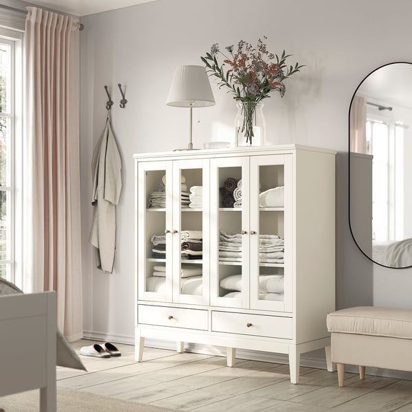 IDANÄS white cabinet, glass doors, storing folded towels, near window with pink curtains, lamp, mirror, cozy decor.