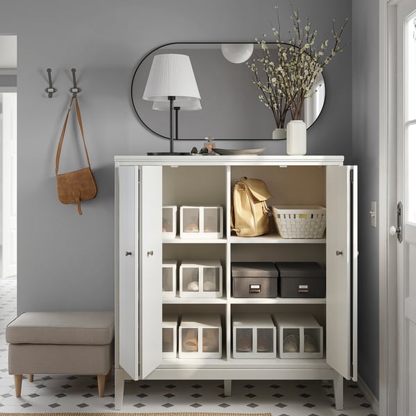 Neat entryway with IDANÄS white cabinet, mirror, lamp, and gray walls. Organized with storage bins, backpacks, flowers, and hooks with bag.