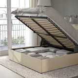 Ottoman bed with slatted base lifted, revealing under-bed storage with folded items. Gray non-woven floor protection, beige upholstery, and headboard.