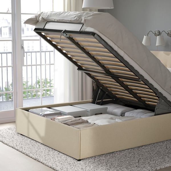 Bed with open storage compartment under mattress, revealing folded linens and pillows, and a slatted base with a metal lift mechanism.