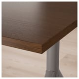 Close-up of IDÅSEN desk top showing dark wood grain pattern, smooth finish, and sturdy metal leg support.