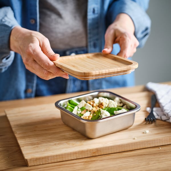 Person placing a bamboo lid on a stainless steel food container filled with salad on a wooden cutting board.