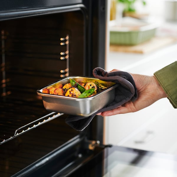 A hand places a metal food container with vegetables into an oven. The container is used for storing leftovers and reheating food.