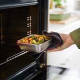 A hand places a metal food container with vegetables into an oven. The container is used for storing leftovers and reheating food.