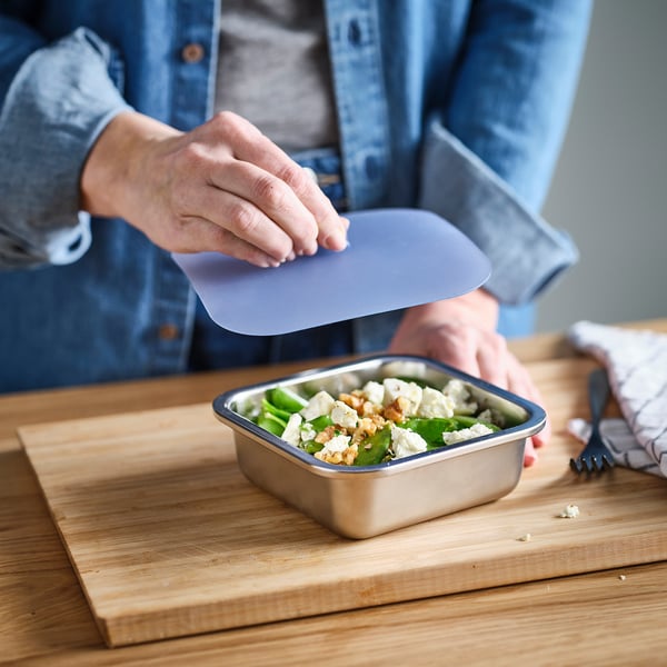 Person places transparent lid on a stainless steel container filled with salad on a wooden table.