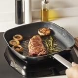 A black grill pan on a stove with steak, mushrooms, and herbs cooking. The non-stick surface ensures easy cooking and cleaning.