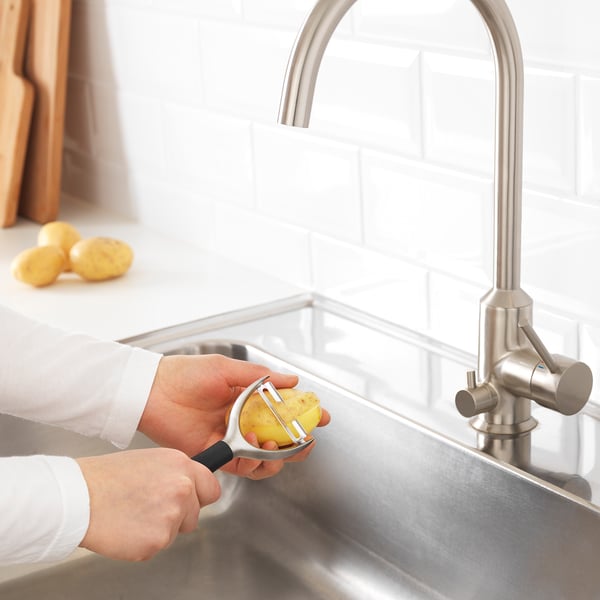 Hand peeling potato over sink with IKEA peeler. Stainless faucet and countertop visible.