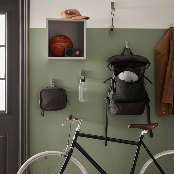 A green wall in a hall displays a bike, backpack, coat, and a water bottle with a strap.