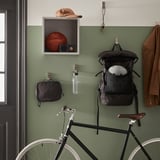 A green wall in a hall displays a bike, backpack, coat, and a water bottle with a strap.