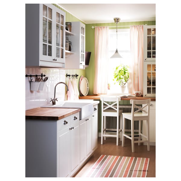 Bright kitchen with white and green cabinets, striped curtains, wooden countertops, and two white stools by the window.