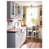 Bright kitchen with white and green cabinets, striped curtains, wooden countertops, and two white stools by the window.