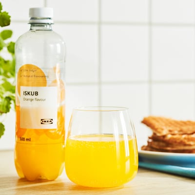A bottle of orange-flavored ikea drink sits next to a glass filled with the beverage on a wooden table.