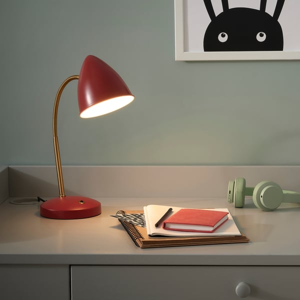 Red ISNÅLEN desk lamp with flexible neck and shade. Near books and headphones, it offers focused light.