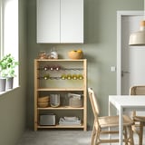 Modern kitchen corner with IVAR pine shelf. Holds jars, plates, and wine. Next to white table and wicker chair. Green wall and white cabinet.