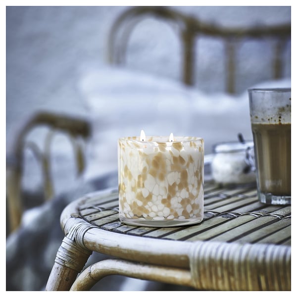 A lit beige and white spotted candle in a glass jar sits on a wicker table, accompanied by a mug.