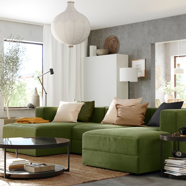 Modern living room with green JÄTTEBO modular sofa, large cushions, round coffee table, pendant lamp, and large window with white curtains.