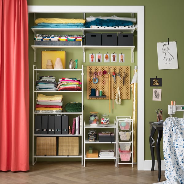 Organized craft room with green walls, white JONAXEL storage shelves, colorful fabrics, and art supplies. Red curtain on left.
