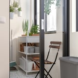 A small, white, metal storage shelf in a room with a balcony door. It holds potted plants and cleaning supplies, with a red chair nearby.
