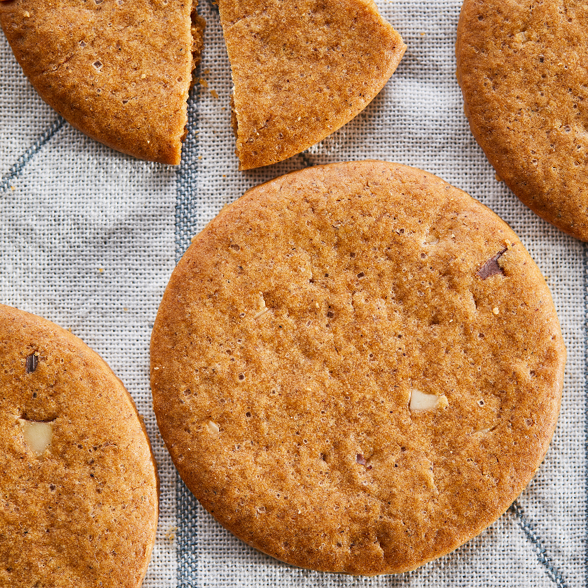 Six golden-brown KAFFEREP ginger biscuits, round, crisp, and speckled with tiny almond crumbs, reposing on a light gray cloth.