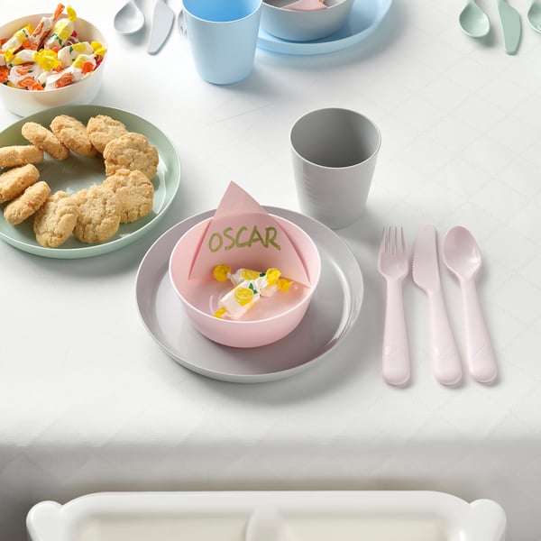 A kids table setting with colorful dishes, cutlery, and snacks. Plates are stacked, and a small bowl holds a napkin with the name oscar on it.