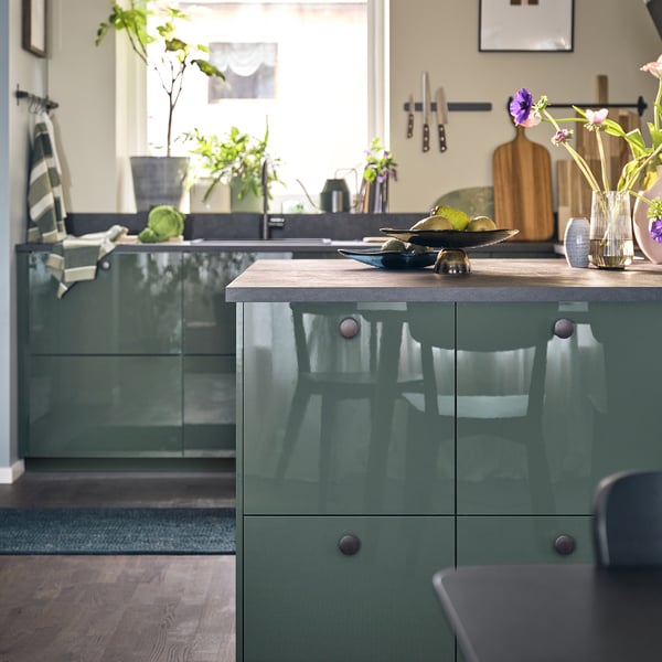 Modern kitchen with glossy green cabinets, wooden counter, decorative items, plants, and natural lighting.