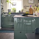 Modern kitchen with glossy green cabinets, wooden counter, decorative items, plants, and natural lighting.