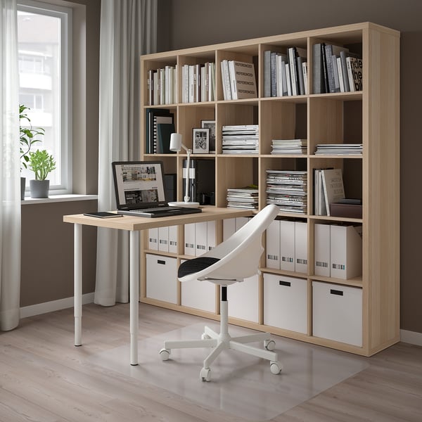 Home office with KALLAX storage. Light wood shelves, white chair. Laptop on simple desk. Organized books and files. Bright, airy room.