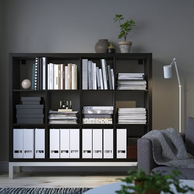 A KALLAX black bookshelf holds books, boxes, and plants; corner of a room with a gray couch and floor lamp.