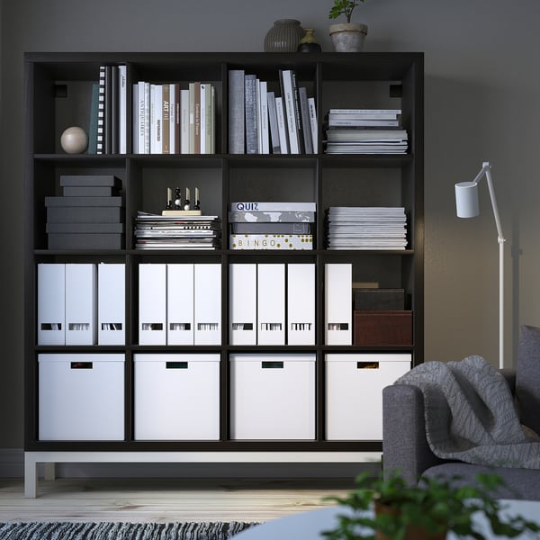 KALLAX black bookshelf. Tall, wide, with multiple compartments. Books, files, and boxes neatly organized. Grey couch and light beside it.