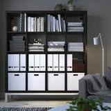 KALLAX black bookshelf. Tall, wide, with multiple compartments. Books, files, and boxes neatly organized. Grey couch and light beside it.