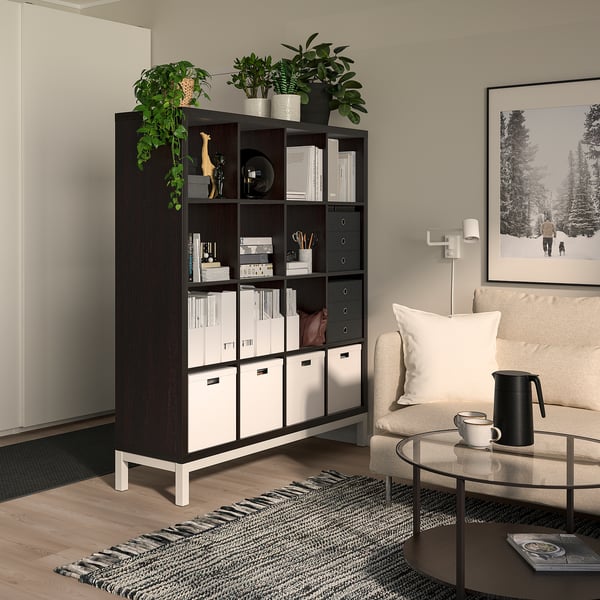 KALLAX shelf with plants, books, decor. Tall divider beside sofa. Cozy room with wooden floors.