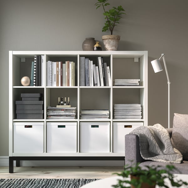 The KALLAX white shelf holds books and decor. Its large size fits many items, prominently displayed against a wall.