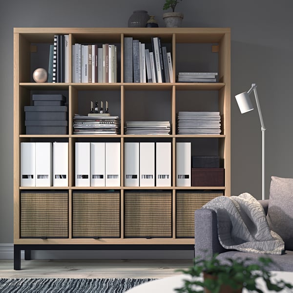 KALLAX shelf with books, files, decor. Tall, next to gray chair, dark gray walls.