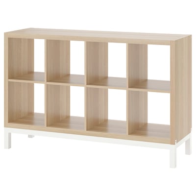 An empty, light wooden KALLAX shelving unit with white legs, featuring multiple cube shelves.