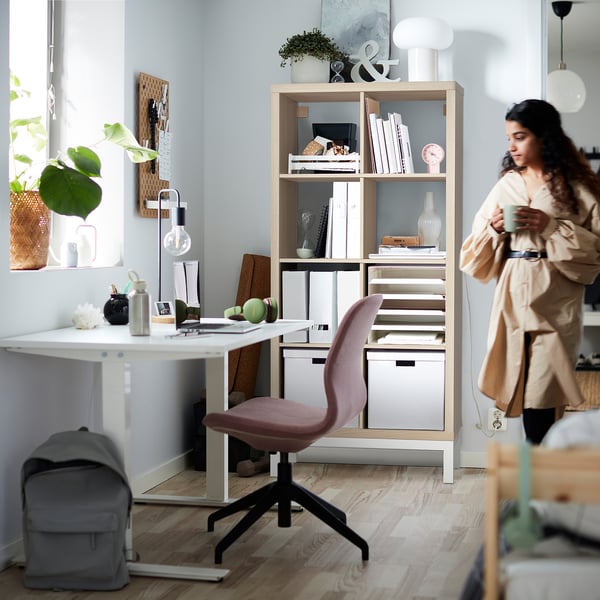 A woman stands by a light wooden KALLAX shelving unit with pink desk chair, holding a cup of coffee in a bright, organized room.