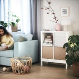 Woman reads on light blue sofa by KALLAX shelf with yarn basket, books, and plant.