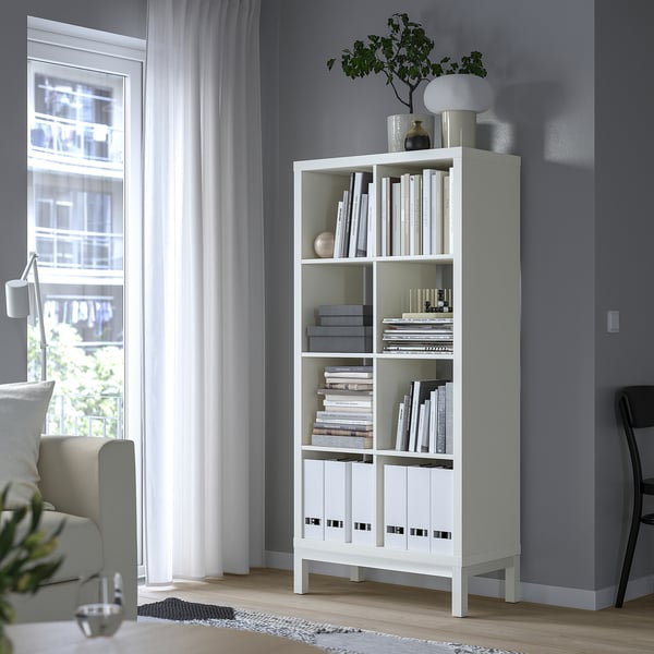 White KALLAX shelves by window, five shelves with books, larger than chair and curtains.