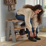 A person tying shoes near a KALLSÖ bench with shoe storage, holding several pairs of footwear, demonstrating its storage function.